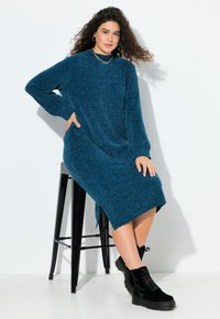 Abito teal a maniche lunghe realizzato in materiale testurizzato, con fessure laterali. Il modello indossa stivaletti neri con lacci, seduto su uno sgabello nero.