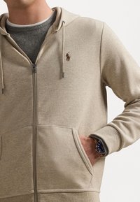 Beige zip-up hoodie i mjukt tyg, med känguruficka och logobrodyr. Underlagret är grått med en ribbad krage.