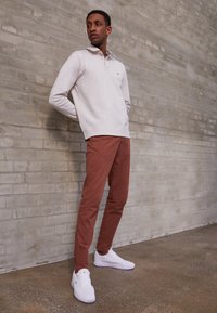 Vaalean beige neule, jossa on kaulus, yhdistettynä ruosteenvärisiin housuihin ja valkoisiin tennareihin. Taustalla on teksturoitu seinä.