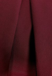 Bordowy materiał o teksturowanym splocie, prezentujący skośne wzory i gładkie wykończenie. Dwa fragmenty zwisają ciasno obok siebie.