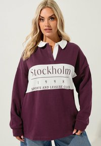 Överdimensionerad långärmad polo i djup lila med vita detaljer. Har en krage och en framgrafik med texten "Stockholm Sports and Leisure Club."
