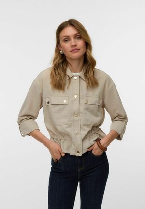 Žena s vlnitými blond vlasmi, oblečená v béžovej gombíkovej bunde so zrolovanými rukávmi a tmavých džínsoch, ruky vreckách na jednoliatom pozadí.