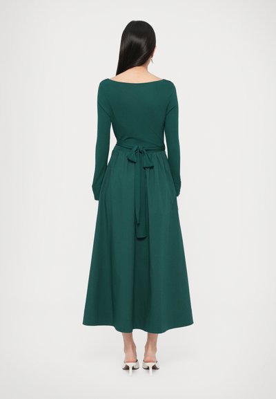 Πράσινο φόρεμα με μακριά μανίκια, ανοιχτή πλάτη σε σχήμα λαιμού, ζώνη δετή στη μέση, ρέουσα φούστα και λεία υφή, συνδυασμένο με λευκά παπούτσια με τακούνι.