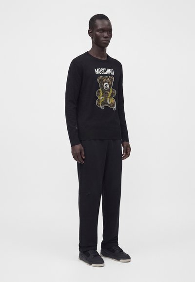 Homme portant un pull Moschino noir avec un motif d'ours en peluche, associé à un pantalon noir et des baskets noires, debout sur un fond blanc uni.