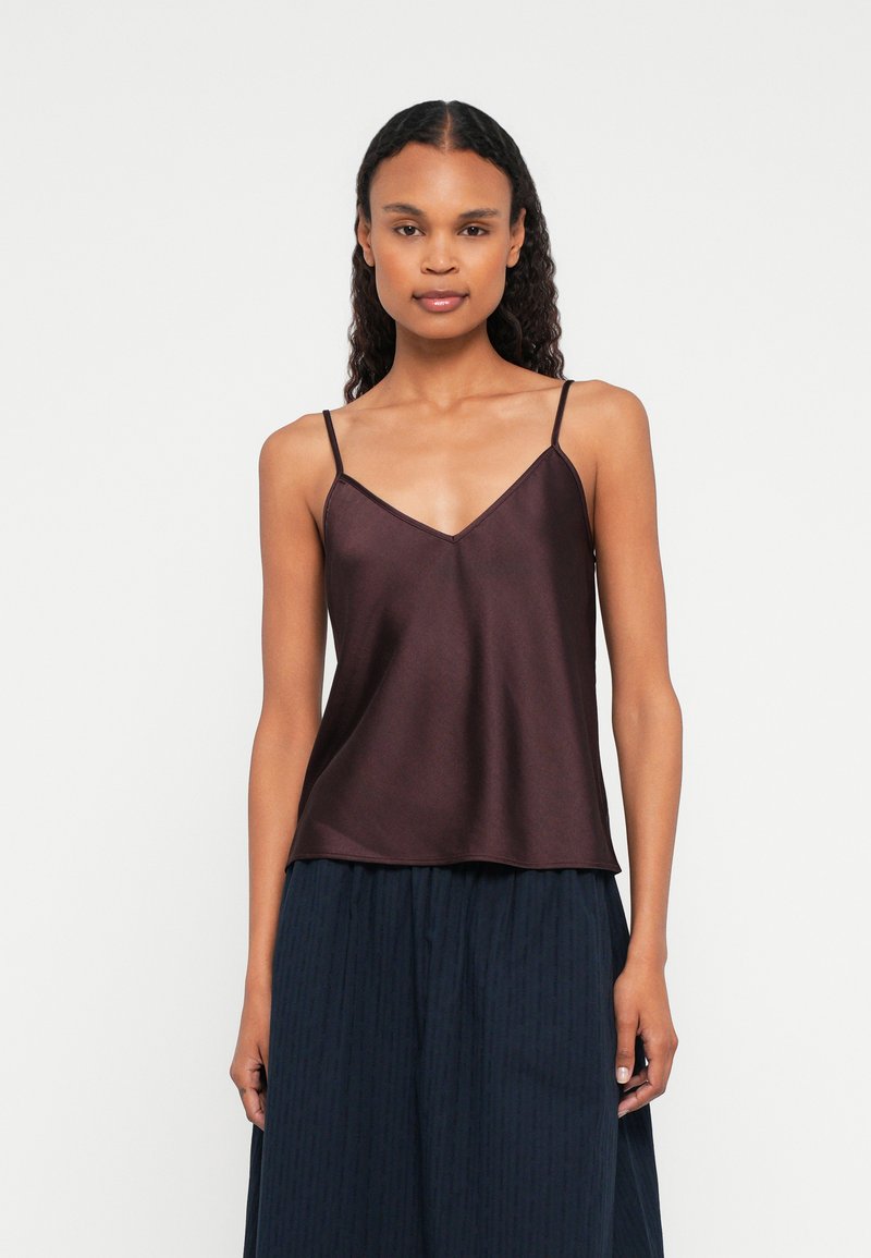 Kobieta z długimi kręconymi włosami nosi ciemnobrązowy satynowy top na cienkich ramiączkach oraz granatową plisowaną spódnicę na jednolitym tle.