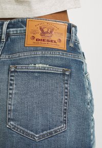 Blå denimjeans har en slit-och-släng bakficka och en brun läderetikett med präglad text och en logotyp vid midjan.