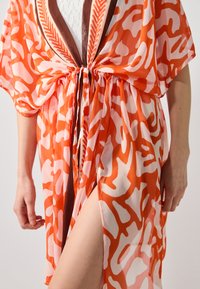 Kimono arancione e bianco con motivo, tessuto trasparente, maniche larghe, vita con cordoncino e accenti marroni, con uno spacco fino alla coscia.