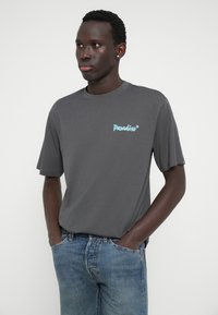Graues Baumwoll-T-Shirt mit kurzen Ärmeln und Rundhalsausschnitt. Verfügt über ein blaues "Paradiso*"-Logo auf der linken Brust. Wird mit blauen Jeans getragen.