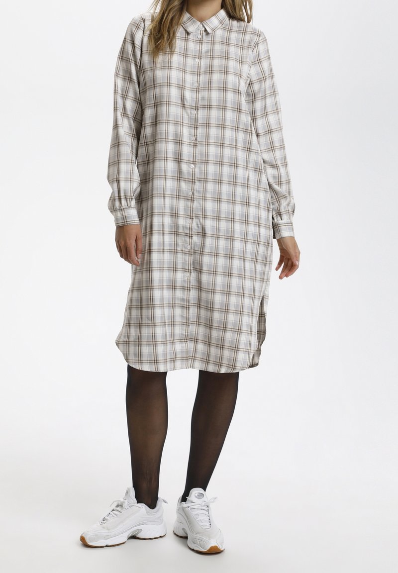 Robe à manches longues en tartan dans des tons beiges et bruns neutres, dotée d'un boutonnage à l'avant et de fentes sur les côtés, portée avec des baskets blanches et des collants noirs.