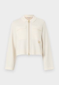 Κομμένο μπουφάν με φερμουάρ σε κρέμα, φτιαγμένο από μαλακό ύφασμα. Διαθέτει γιακά, δύο τσέπες στο στήθος, χρυσό φερμουάρ και κυκλικό διακοσμητικό στοιχείο.