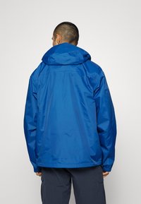 Chaqueta impermeable azul con capucha, que presenta puños elásticos y un corte holgado. Textura suave y diseño minimalista, adecuada para usar al aire libre.