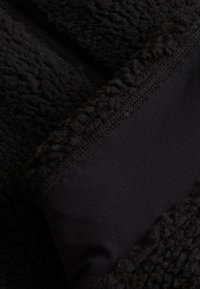 Svart fleece-tyg med en texturerad yta, som har en kontrast av slät svart material vid kanten, vilket framhäver sömnadsdetaljer.