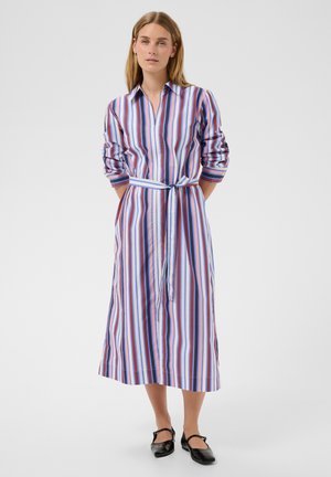 Femme aux cheveux lisses portant une robe à manches longues ceinturée avec des rayures verticales bleues, roses et blanches, et des chaussures plates noires, debout les mains dans le dos.