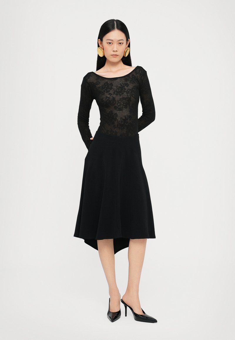 Abito nero con un corpino in pizzo trasparente, maniche lunghe e una gonna svasata con orlo asimmetrico. Abbinato a tacchi neri a punta.