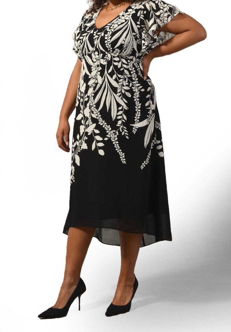 Frau trägt ein schwarzes Midikleid mit weißen Blumenmustern, kurzen Rüschenärmeln und schwarzen High Heels, steht mit einer Hand in der Hüfte.