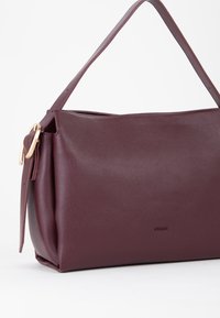 Burgunder lædertaske med en glat, struktureret overflade. Har en enkelt bred rem og detaljer i guldfarvet metal på siden.
