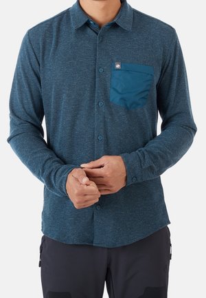 Mann trägt ein blaues Hemd mit langen Ärmeln und kontrastierender Brusttasche, das erknöpft ist, und richtet die Ärmelmanschetten vor einem einfarbigen Hintergrund.