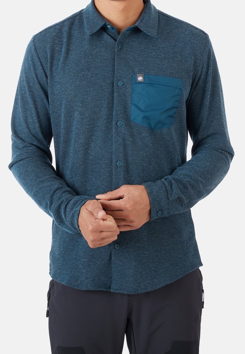 Homme portant une chemise bleue à manches longues boutonnée avec une poche poitrine contrastante, ajustant les poignets des manches sur un fond uni.