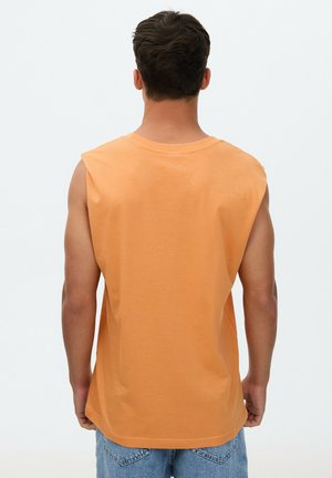 Hombre con cabello corto y oscuro, que lleva una camiseta sin mangas naranja y vaqueros azules, de pie de espaldas contra un fondo liso.