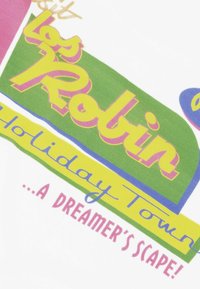 Camiseta blanca con texto colorido: "Les Robin Holiday Town" en amarillo y rosa, con detalles en verde y azul y un diseño de fuente juguetón.