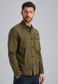 Camicia verde oliva con bottoni a pressione, due tasche sul petto, bottoni neri e un'etichetta in tessuto. Realizzata in materiale testurizzato, con maniche lunghe.