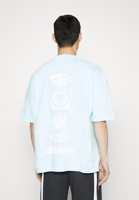 Světle modré tričko s krátkým rukávem a kulatým výstřihem ve stylu tie-dye. Na zadní straně má bílé grafické texty, včetně log a vzorů.