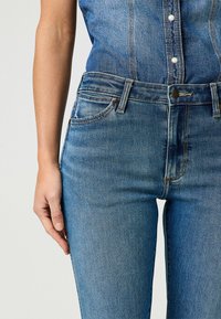 Jeans de mezclilla azul desgastados con un diseño de tiro medio, cinco bolsillos y costuras en contraste, que cuentan con una cremallera y un cierre de botón metálico.