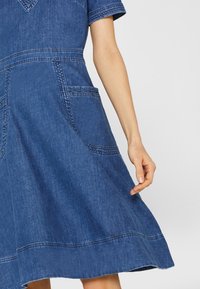 Teepparin denim, joka on varustettu laajentuvalla siluetilla, lyhyillä hihoilla, viistoin taskuin, kontrastivärillä ommelluilla saumoilla ja syvän sinisellä värillä.