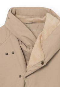 Béžová puffová bunda so žiarivým fleece golierom v krémovej farbe. Obsahuje dvojradové zlaté gombíky a preložený dizajn pre väčšie teplo.