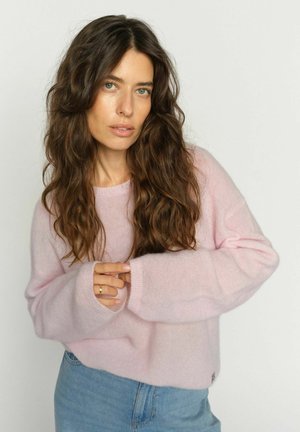 Frau mit langen, welligen braunen Haaren, die einen hellrosa Pullover und blaue Jeans trägt, steht und schaut mit verschränkten Händen in die Kamera.