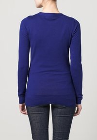 Långärmad lila tröja med rund halsringning, ribbade muddar och fåll, figursydd modell och slät textur. Buren över mörka jeans.