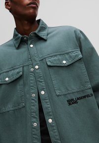 Žalias džinsinis marškinis su apykakle, dviem krūtinės kišenėmis, sidabriniais snapeliais ir išsiuvinėtu "KARL LAGERFELD JEANS" užrašu.