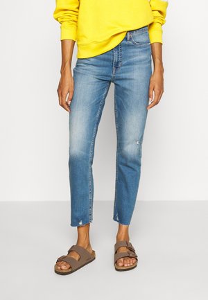 Personne portant un jean court bleu délavé, un haut jaune à manches longues et des sandales marron à double bride, debout devant un fond uni.