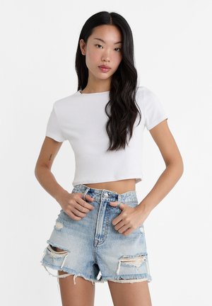 Junge Frau mit langen dunklen Haaren, die ein weißes, kurzes T-Shirt und hoch taillierte, ausgefranste Denim-Shorts trägt.