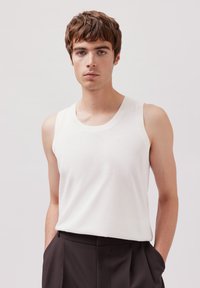 Weißes ärmelloses Tanktop aus Baumwolle mit geripptem Ausschnitt, taillierter Form und strukturiertem Stoff. Kombiniert mit dunklen Hosen.