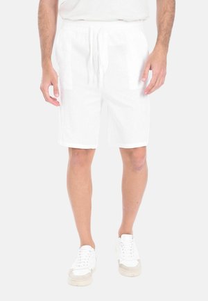 Uomo che indossa pantaloncini bianchi con coulisse e scarpe da ginnastica bianche, in piedi contro uno sfondo chiaro e uniforme.