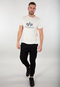 Hvid t-shirt med korte ærmer og et blåt grafisk logo på fronten, parret med sorte cargobukser og sorte sneakers. Ensfarvet baggrund.