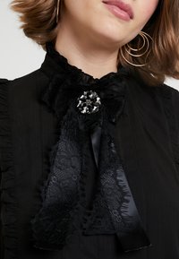 Černá halenka s krajkovým límcem s volánky, saténovou stuhou a ozdobnou broží zdobenou černými drahokamy.