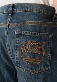 Jeans de mezclilla azules con un gran logo de árbol naranja bordado en el bolsillo trasero, con costura estándar y cierre de botón.