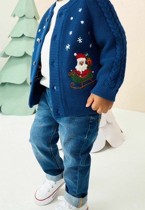 Enfant portant un cardigan bleu tricoté avec traîneau du Père Noël et flocons de neige, jean, baskets blanches, debout près d'arbres décoratifs verts et blancs.