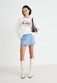 Sweatshirt blanc avec le texte "hello" rouge, jupe en denim bleu clair et bottes crème montantes à bouts pointus. Sac marron, accessoires minimalistes.