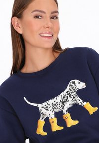 Tengerészkék pulóver dalmata mintával, amely sárga gumicsizmát visel. A pulóver kerek nyakú és hosszú ujjú, puha tapintású anyagból.