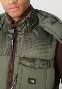 Green padded vest with a textured upper and smooth lower section. Features a zip closure, front pocket, and branded label.
