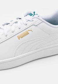 Hvide lædersneakers med en glat overflade, udstyret med gyldne og teal accenter, samt en hvid gummisål. Synlig brandlogo på siden.