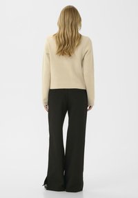 Beige kötött pulóver hosszú ujjakkal, fekete, széles szárú nadrággal párosítva, amelynek alján hasítékok vannak. Simaszerkezet, minimalista dizájn.