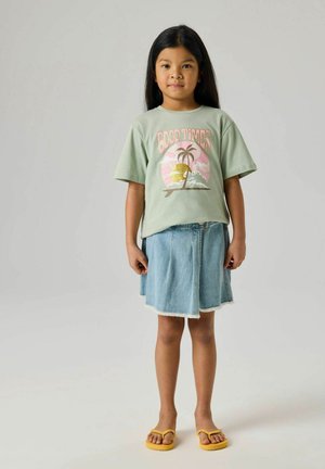 Young girl standing wearing green "Good Times" t-shirt, denim skirt, and yellow flip-flops against plain light background.