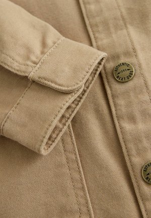 Hellbraunes Hemd aus Stoff mit einem umgeschlagenen Ärmelaufschlag, sichtbaren Nähten und einem runden Messingknopf mit der Aufschrift "Authentic 1982 Next Dude".