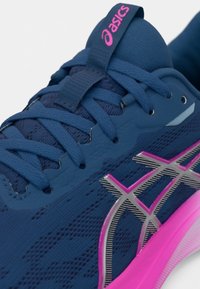 Μπλε αθλητικό παπούτσι με υφή στο πάνω μέρος, ροζ και ασημί λεπτομέρειες στον λογότυπο και αυλάκωση στο τακούνι για καλύτερη πρόσφυση. Οι κορδόνια είναι επίπεδα και υφασμένα.