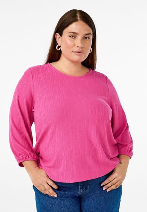 Pink long-sleeve top made of textured fabric, featuring a round neckline and gathered cuffs, paired with blue denim jeans.