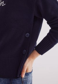 Marineblauwe gebreide trui met geribbelde manchetten en zoom, voorzien van een zijknopenlijst en een gestructureerde afwerking. Casual fit, gedragen met denim.